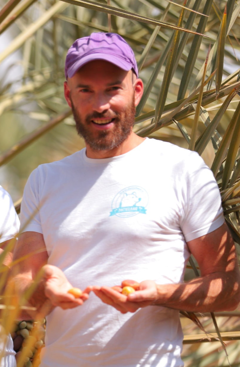 Ein bärtiger Mann mit weißem T-Shirt und lila Kappe steht zwischen Palmwedeln, lächelt und hält mehrere kleine gelbe Früchte in den Händen.